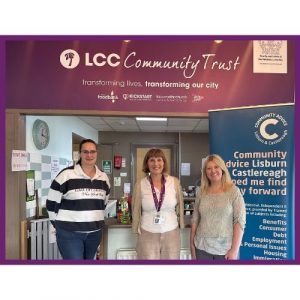 Three people stand inside LCC Community Trust Inspire Café beside a Community Advice Lisburn & Castlereagh banner.